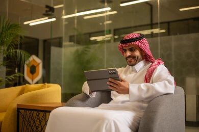 استخدام الاجهزة التقنية الحديثة، ادارة مهام العمل اليومية، رجل عربي خليجي سعودي يرتدي الزي التقليدي والشماغ يجلس في مكتب يحمل بيده جهاز التابلت، مهن ووظائف سعودية