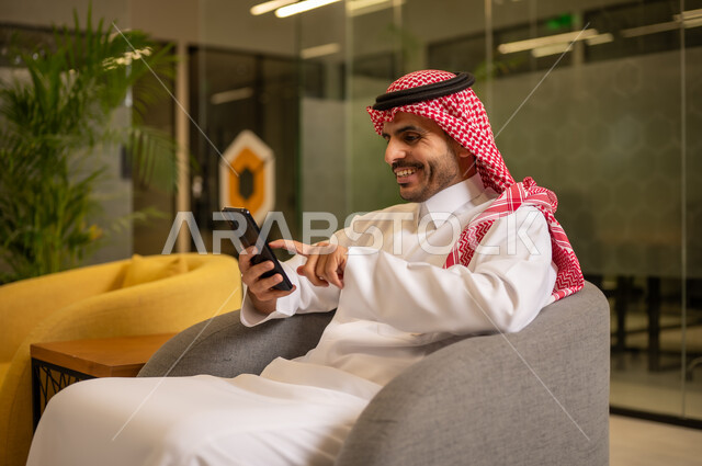 إيماءات الجودة والرضا والقبول, رجل عربي خليجي سعودي مبتسم يرتدي الثوب التقليدي والشماغ يقوم بإجراء مكالمة هاتفية عبر الهاتف المحمول, العمل في المجال المكتبي, متابعه الأعمال الادارية