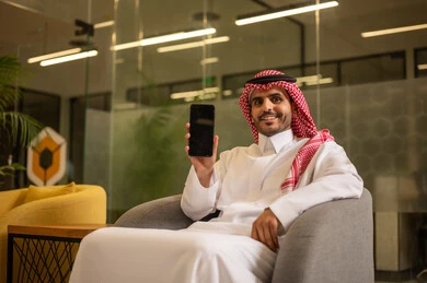 Follow-up on administrative tasks, gestures of quality, satisfaction, and acceptance. A smiling Arab Gulf Saudi man wearing traditional attire and a ghutrah is making a phone call via a mobile phone, working in an office environment.