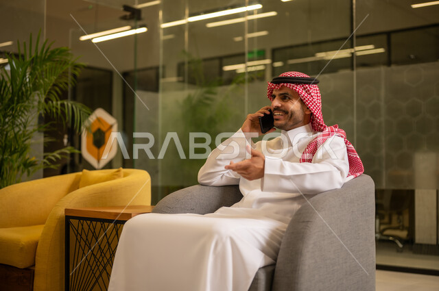 العمل في المجال المكتبي, إيماءات الجودة والرضا والقبول, رجل عربي خليجي سعودي مبتسم يرتدي الثوب التقليدي والشماغ يقوم بإجراء مكالمة هاتفية عبر الهاتف المحمول,متابعه الأعمال الادارية