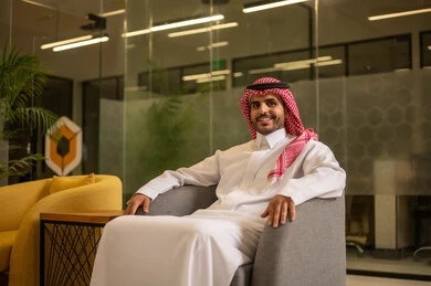 Expressions of joy and happiness, a smiling Arab Gulf Saudi man wearing traditional attire and a ghutrah looking at the camera, working in an office environment.