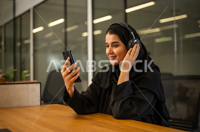 الإجابة على استفسارات العملاء, موظفة عربية خليجية سعودية مبتسمة ترتدي العباءة والحجاب تضع سماعات الرأس (الهيدفون) تستخدم الهاتف المحمول، العمل في المجال المكتبي