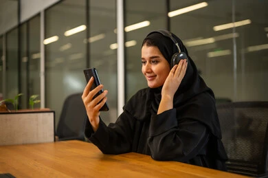 الإجابة على استفسارات العملاء, موظفة عربية خليجية سعودية مبتسمة ترتدي العباءة والحجاب تضع سماعات الرأس (الهيدفون) تستخدم الهاتف المحمول، العمل في المجال المكتبي