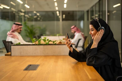 Business follow-up and development, answering customer inquiries, a smiling Arab Gulf Saudi female employee wearing an abaya and hijab, wearing headphones, using a mobile phone, working in an office environment.