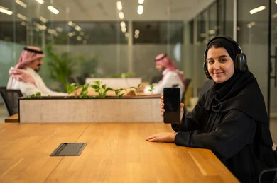 عرض شاشة سوداء فارغة, متابعة وتطوير الأعمال، الإجابة على استفسارات العملاء, موظفة عربية خليجية سعودية مبتسمة ترتدي العباءة والحجاب تضع سماعات الرأس (الهيدفون) تستخدم الهاتف المحمول، العمل في المجال المكتبي