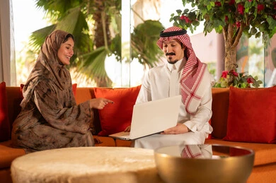 Saudi Couple in Traditional Attire Using Laptop Indoors
