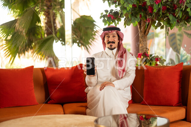 Saudi Man in Thobe Holding Smartphone on Sofa