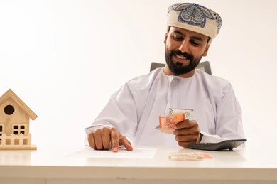Omani Man Counting Money with House Model on Desk