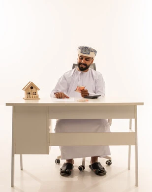 Omani Man Counting Money with House Model on Desk