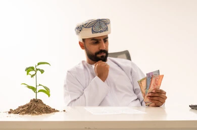 رجل عُماني يمسك أوراقاً نقدية بجانب نبتة نامية