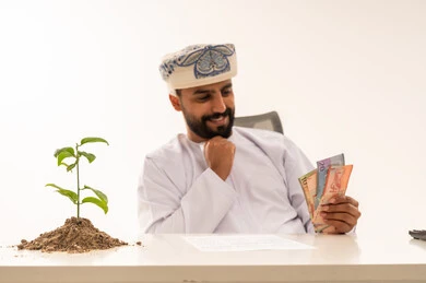 رجل عماني يحمل عملات ورقية بجانب شتلة نمو