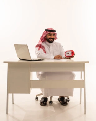 رجل سعودي يجلس خلف مكتب عمل ويمسك ساعة منبه