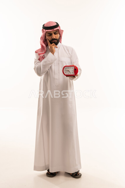 رجل سعودي بالثوب التقليدي يفكر مع ساعة منبه حمراء