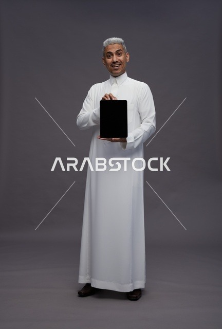 رجل سعودي بالثوب يحمل جهاز لوحي بخلفية رمادية