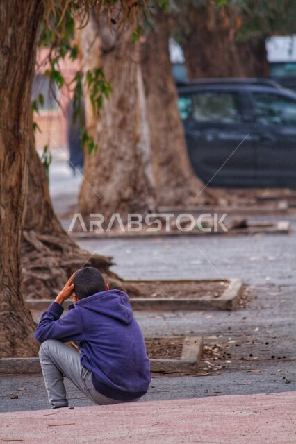 صورة من الخلف لطفل عربي يجلس على جانب الطريق ويضع يده على رأسه، جذوع أشجار كبيرة، سيارة جيب، التفكير والحزن