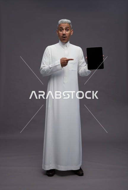رجل سعودي يحمل جهاز لوحي أمام خلفية رمادية