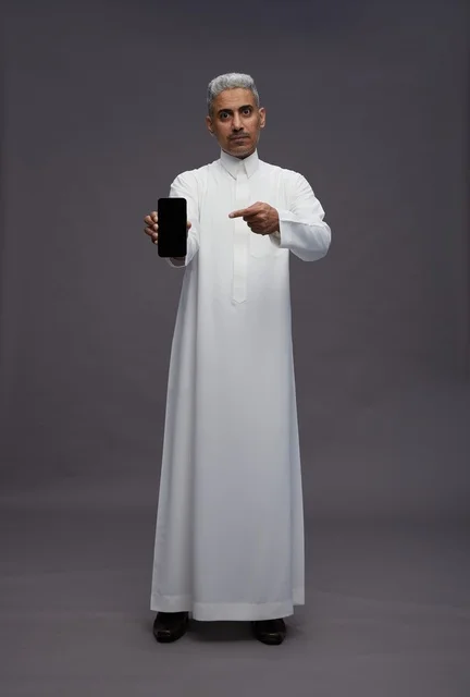 رجل سعودي يشير إلى شاشة هاتف ذكي بخلفية رمادية