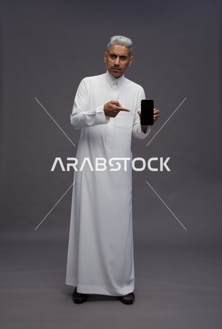 رجل سعودي بالثوب الأبيض يشير إلى شاشة هاتف ذكي