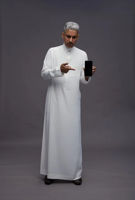 رجل سعودي يرتدي الثوب الأبيض ويحمل هاتفاً ذكياً في الاستوديو