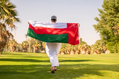 رجل عُماني يحمل العلم الوطني بالزي التقليدي في حديقة