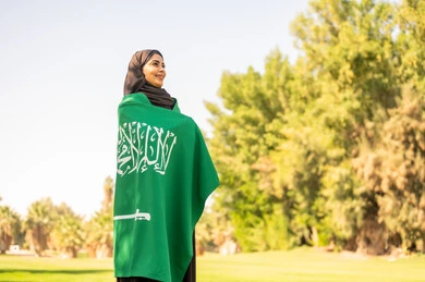 امرأة سعودية تلتف بالعلم الوطني السعودي في حديقة خضراء