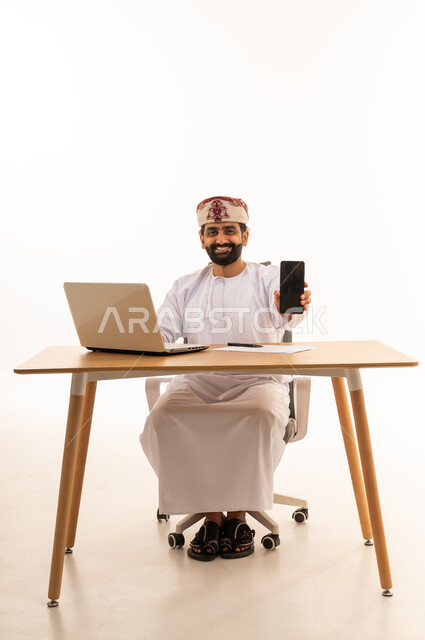 رجل عماني جالس خلف مكتب يحمل هاتف ذكي