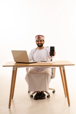 رجل عماني جالس خلف مكتب يحمل هاتف ذكي