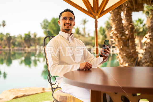 Saudi Man in Thobe Using Smartphone by Lake