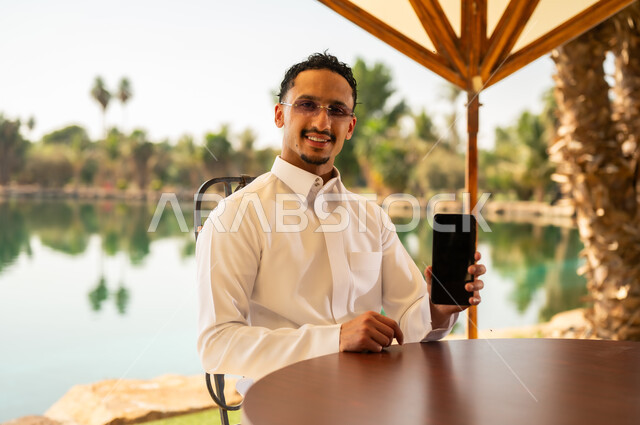 Saudi Man in Thobe Holding Smartphone by Lake