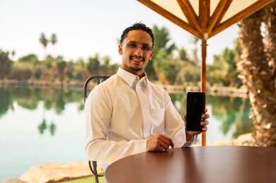 رجل سعودي بالثوب يحمل هاتفاً ذكياً أمام البحيرة