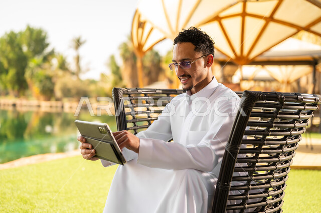 رجل سعودي بالثوب يستخدم جهاز لوحي في الحديقة