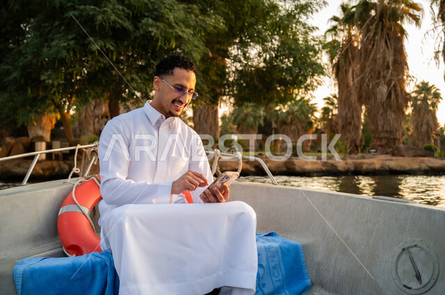 رجل سعودي بالثوب يستخدم الهاتف في قارب وقت الغروب