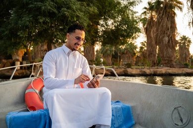 رجل سعودي بالثوب يستخدم الهاتف في قارب وقت الغروب