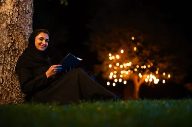 امرأة سعودية تقرأ كتاب في حديقة ليلية مع أضواء بوكيه