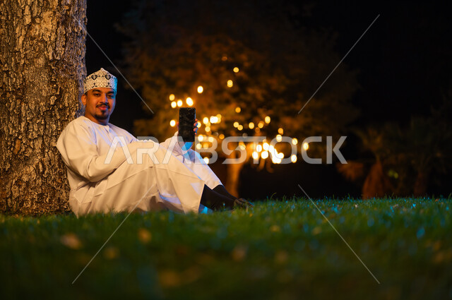 Omani Man Holding Smartphone in Night Park with Lights