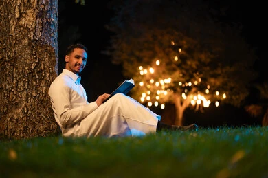 شاب سعودي يقرأ كتاباً تحت شجرة في حديقة ليلية