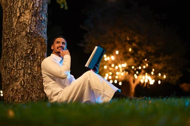 رجل سعودي يرتدي الثوب يقرأ كتاباً في الخارج ليلاً