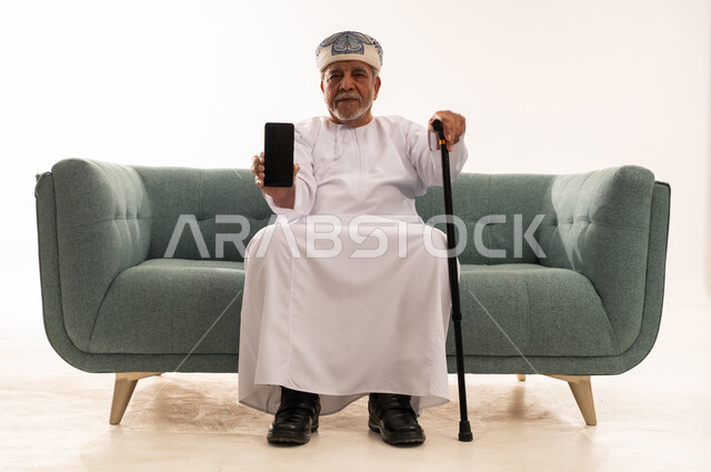 رجل عماني مسن يحمل هاتفاً ذكياً على الأريكة