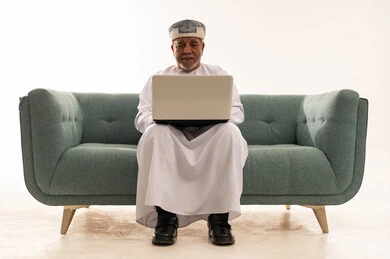 رجل عُماني مسن يستخدم الحاسوب المحمول على أريكة خضراء