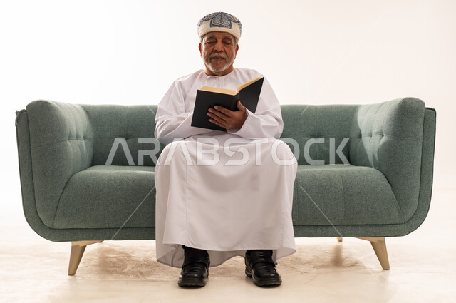 رجل عماني مسن يقرأ كتاباً على الأريكة بخلفية بيضاء