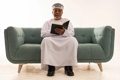 رجل عماني مسن يقرأ كتاباً على الأريكة بخلفية بيضاء