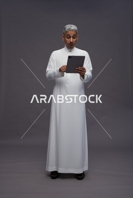 رجل سعودي بالثوب مع جهاز لوحي بخلفية رمادية