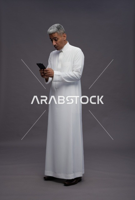 رجل سعودي بالثوب يستخدم الهاتف الذكي بخلفية رمادية