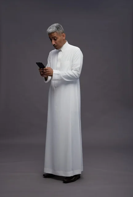 رجل سعودي بالثوب يستخدم الهاتف الذكي بخلفية رمادية