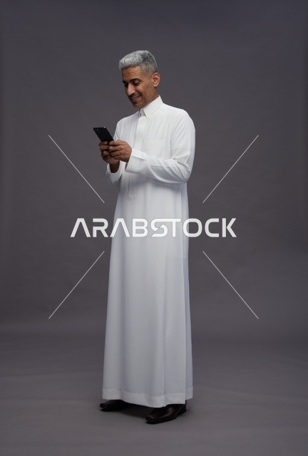 رجل سعودي بالثوب يستخدم هاتفه الذكي بخلفية رمادية