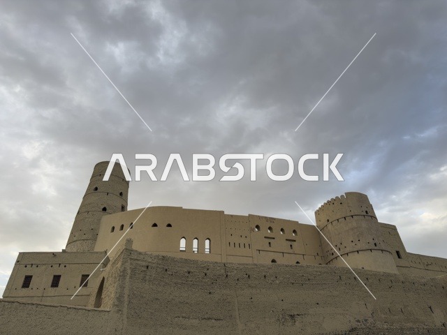 منظر لقلعة بهلا التاريخية في سلطنة عمان يظهر العمارة الطينية العريقة وأبراجها الدفاعية تحت سماء غائمة
