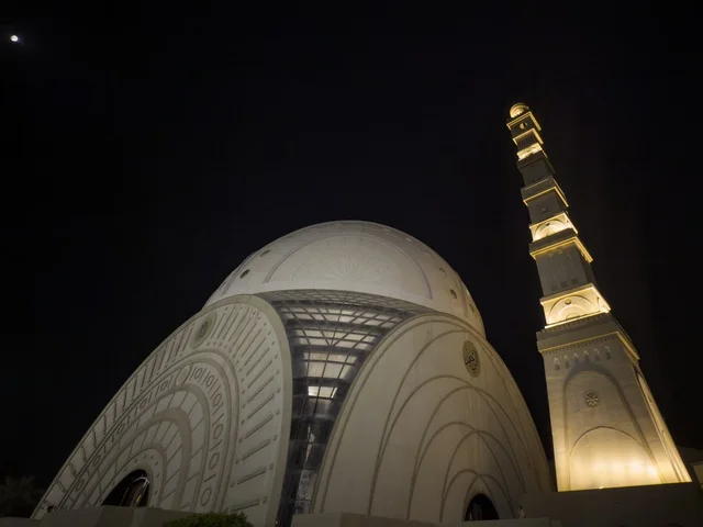 منظر ليلي لجامع الإسلام في مسقط يظهر القبة البيضاء والمئذنة المضيئة في أجواء روحانية هادئة