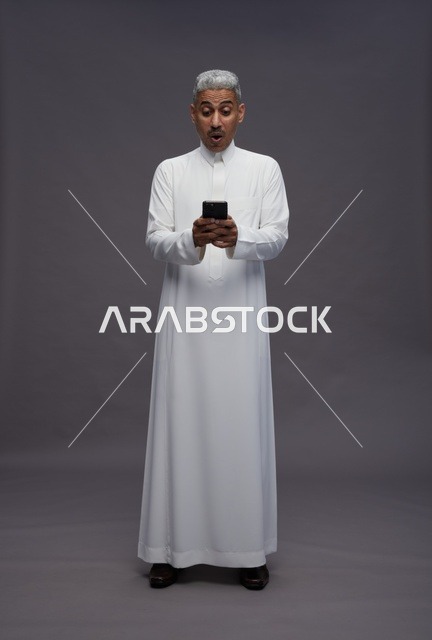 رجل سعودي بالثوب يستخدم هاتفه الذكي بتعبير مندهش