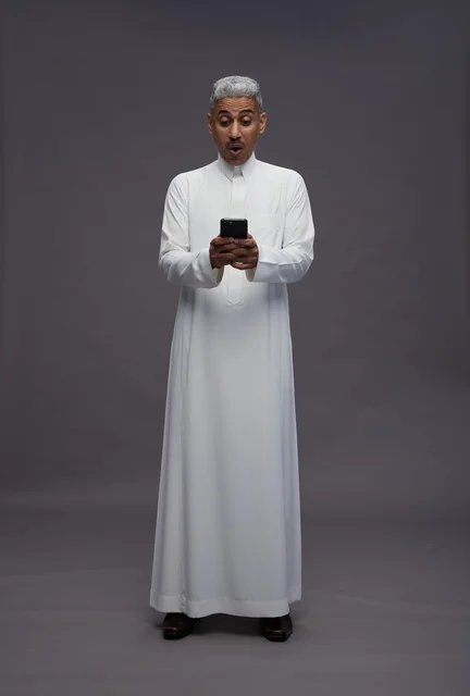 رجل سعودي بالثوب يستخدم هاتفه الذكي بتعبير مندهش