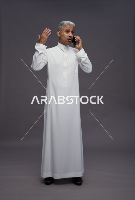 رجل سعودي بالثوب الأبيض يتحدث بالهاتف بخلفية رمادية